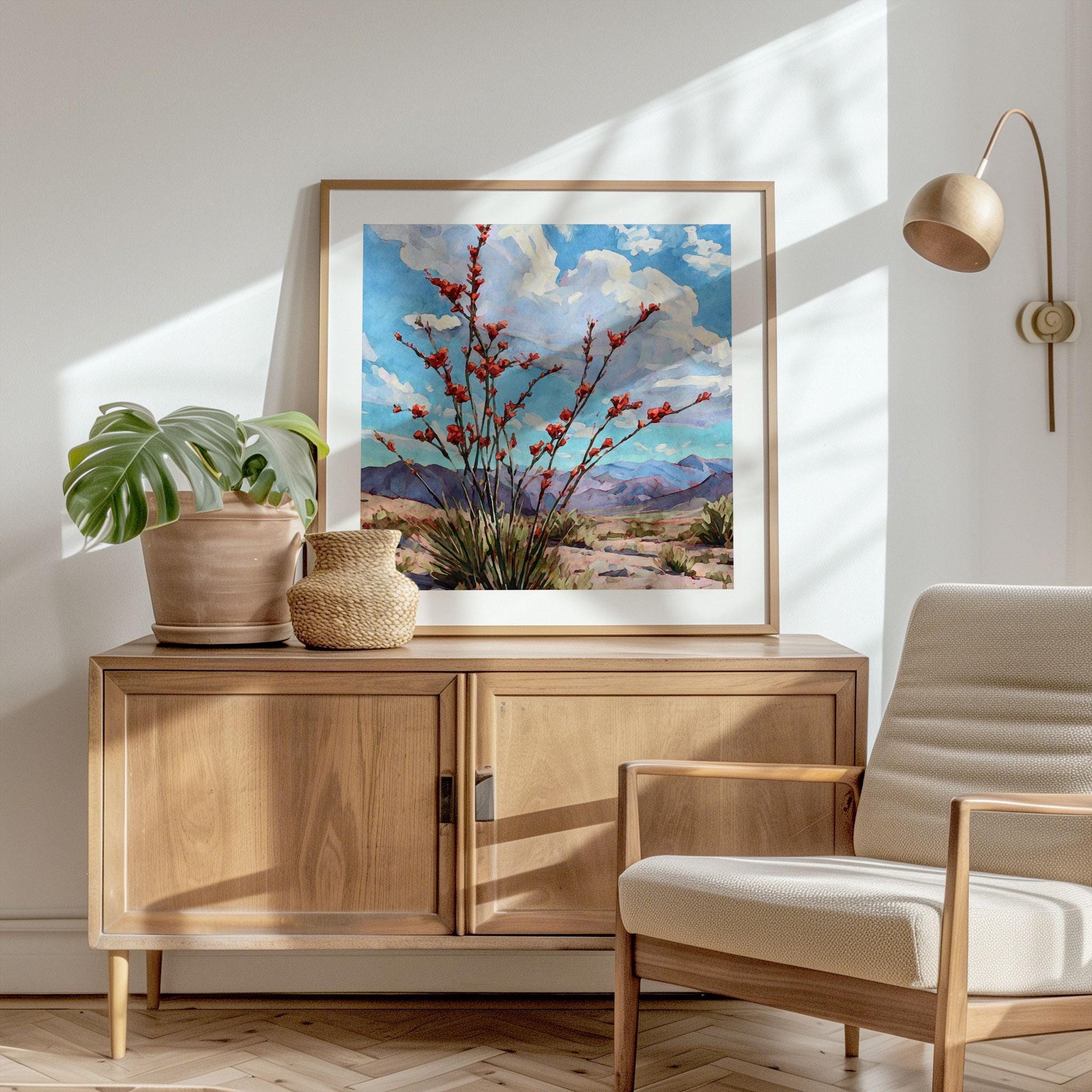 Framed desert landscape painting of a red-flowering ocotillo against blue skies and distant purple mountains, displayed on a light wood credenza. The setting includes a potted monstera, woven vase, and mid-century modern chair under warm natural sunlight, creating a calm, airy, minimalist interior.