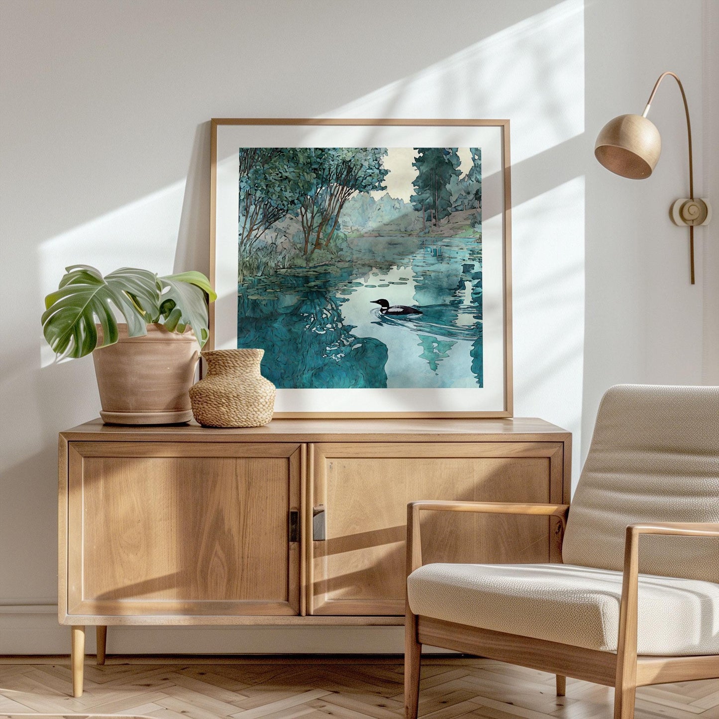 Framed art print of a loon gliding across a reflective lake with trees in the background, styled on top of a wooden sideboard beside a potted plant and woven vase, in a bright modern room with a neutral armchair and wall lamp.