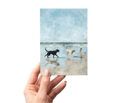 Hand holding a greeting card featuring a watercolor-style illustration of three dogs—one black and two golden—playing on a sandy beach with gentle waves under a bright blue sky.