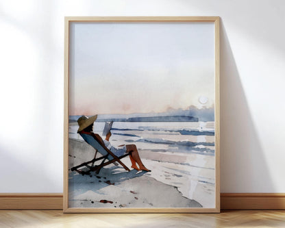 Framed watercolor painting of a woman relaxing on a beach chair, wearing a wide sun hat and reading a book by the shoreline. The soft pastel sky blends into the horizon as gentle waves roll in, creating a serene coastal sunset scene.