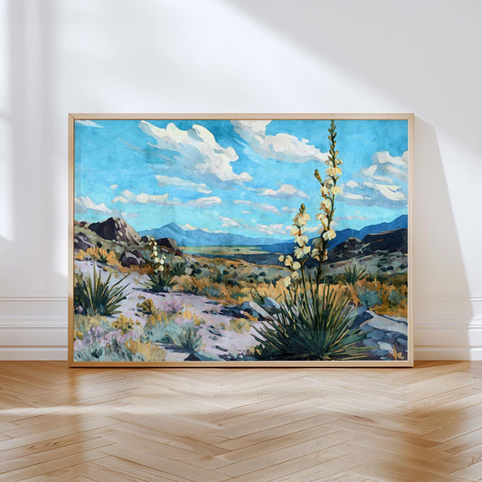 Framed desert landscape painting featuring yucca plants in bloom against a backdrop of rolling hills, rocky terrain, and a bright blue sky with scattered white clouds, displayed leaning against a wall on a wooden floor.