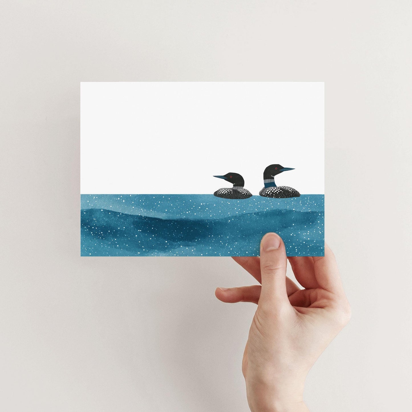 Hand holding a minimalist greeting card featuring two loons illustrated in black and white, floating on deep blue, star-speckled water against a crisp white background.