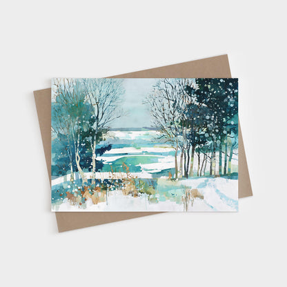 Watercolor greeting card featuring a serene winter landscape with snow-covered fields, bare trees, and evergreens in shades of blue, teal, and white, with soft brushstrokes and splattered paint suggesting falling snow. Card is displayed with a kraft paper envelope.