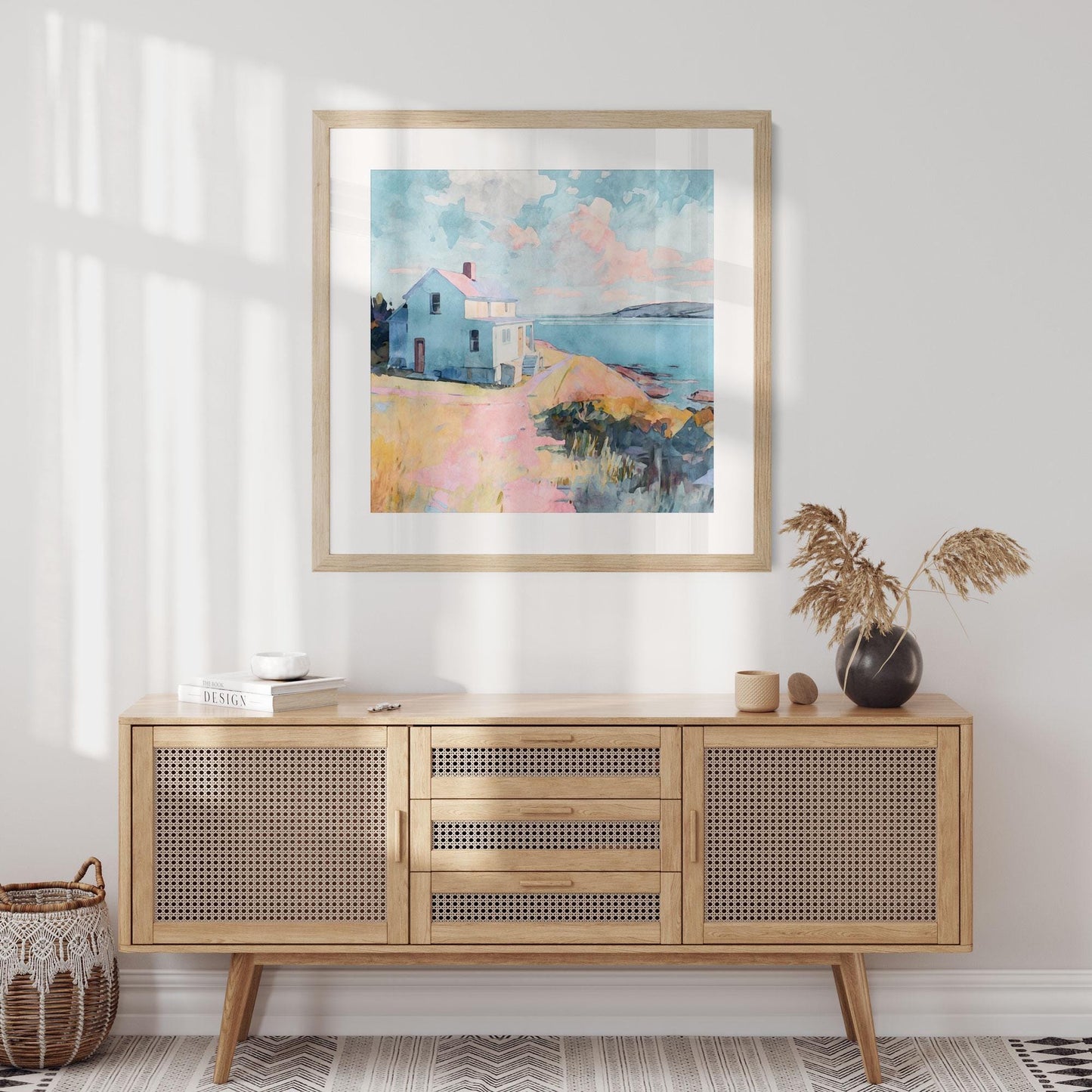 Square framed watercolor painting of a coastal scene with a light blue house on a bluff overlooking the ocean, hung above a light wood cabinet with woven rattan doors. The room is styled with a black vase holding dried grasses, a small basket on the floor, and books on the cabinet, creating a warm, airy, and modern coastal aesthetic.