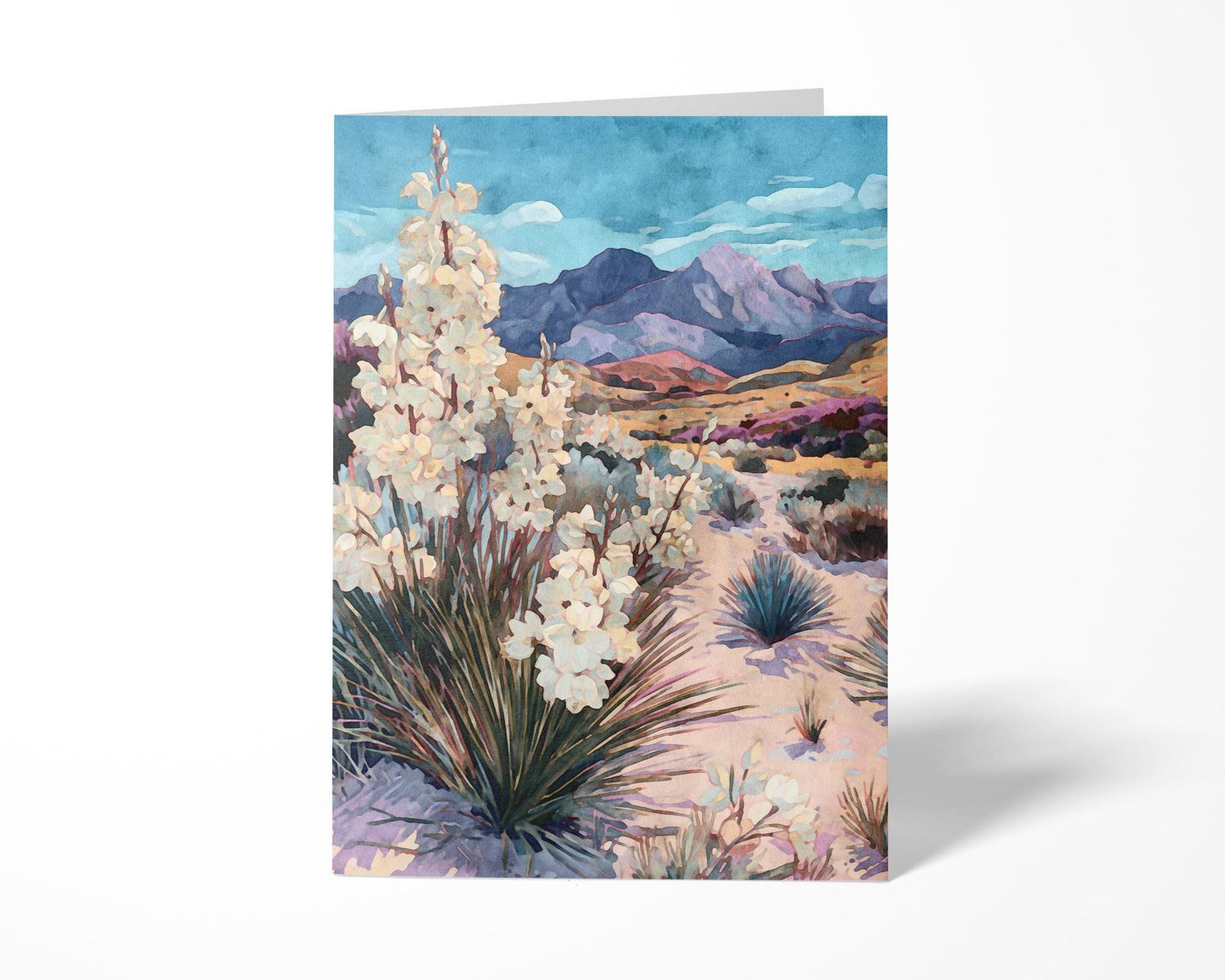 Greeting card featuring a watercolor-style desert scene with blooming yucca plants in the foreground, sandy terrain, and purple mountains beneath a blue sky, displayed standing upright against a white background.