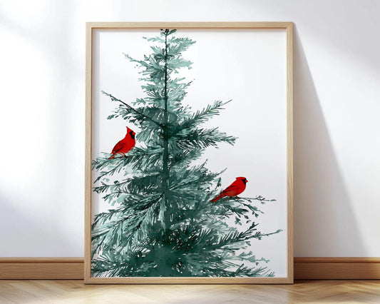 Minimalist watercolor art print of a green pine tree with two bright red cardinals perched on its branches, framed in light wood and leaning against a white wall on a wooden floor.