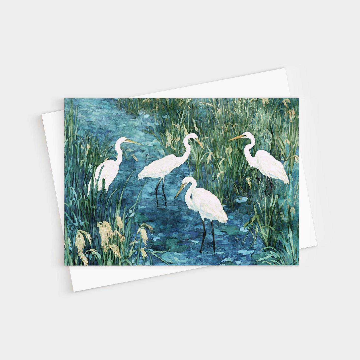 Greeting card featuring a watercolor-style painting of five white egrets standing in a marshy blue-green wetland, surrounded by tall grasses and reeds. Card is paired with a white envelope.