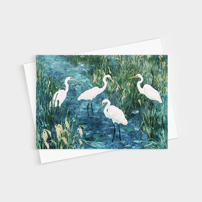 Greeting card featuring a watercolor-style painting of five white egrets standing in a marshy blue-green wetland, surrounded by tall grasses and reeds. Card is paired with a white envelope.