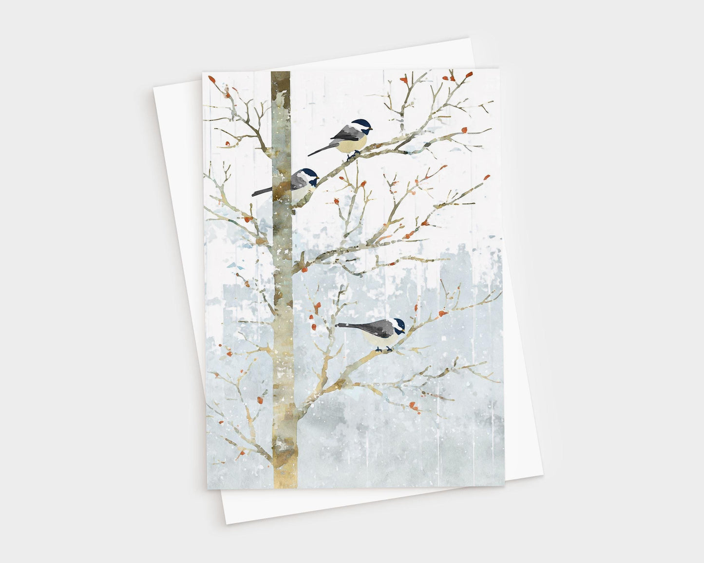 Greeting card featuring a watercolor-style illustration of three chickadees perched on bare winter branches. Soft gray-blue snowy background with scattered orange berries. The card is shown with a white envelope behind it.