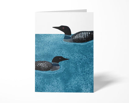 Folded vertical greeting card featuring two loons illustrated in black and white, swimming on rippling textured deep blue water with a clean white background, displayed standing upright.
