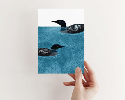 Hand holding a vertical greeting card featuring two loons illustrated in black and white, swimming on textured deep blue water with gentle ripples, set against a white background.