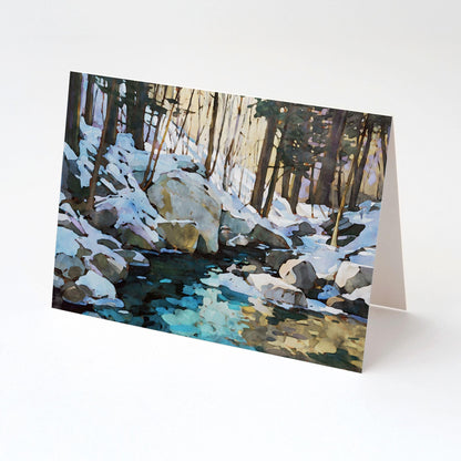 Greeting card standing upright, featuring a watercolor-style snowy forest scene with large rocks and a creek reflecting icy blue and golden tones beneath tall trees.