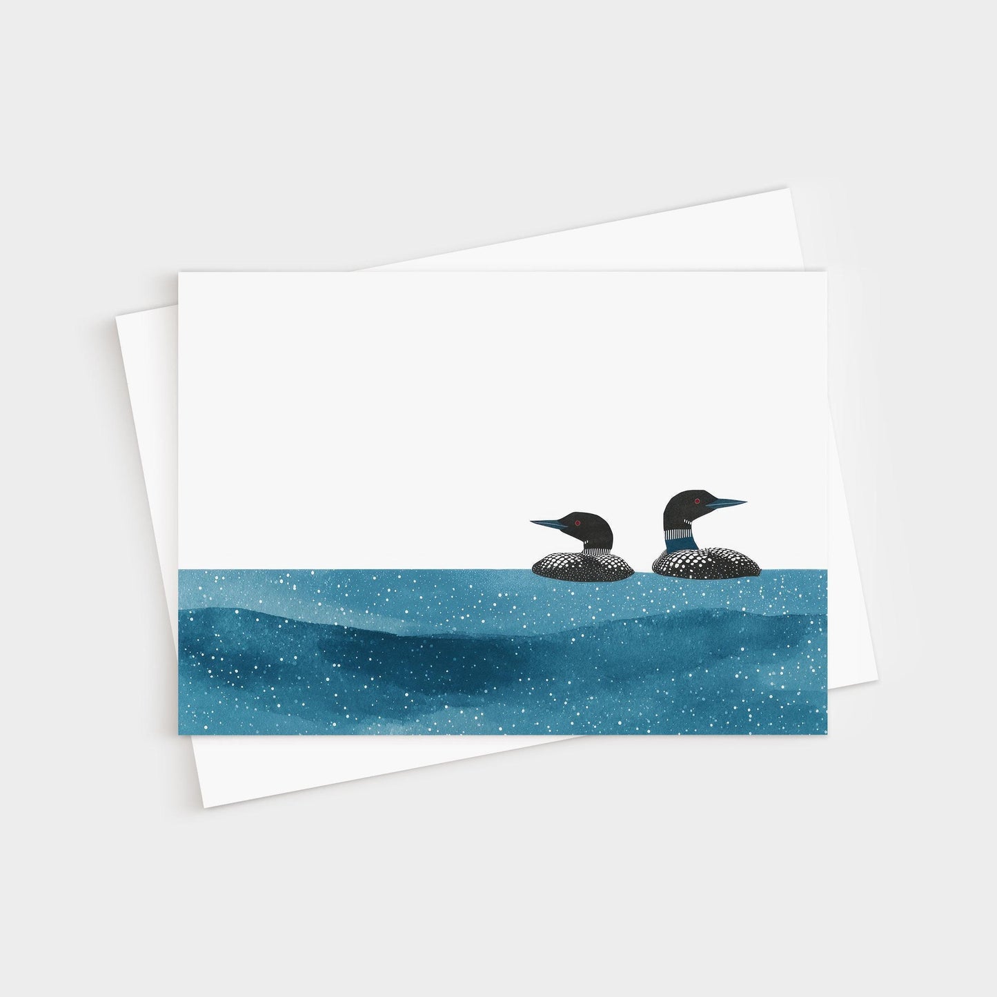 Minimalist greeting card with two loons illustrated in black and white, floating on a deep blue, star-speckled water pattern against a clean white background, paired with a white envelope.