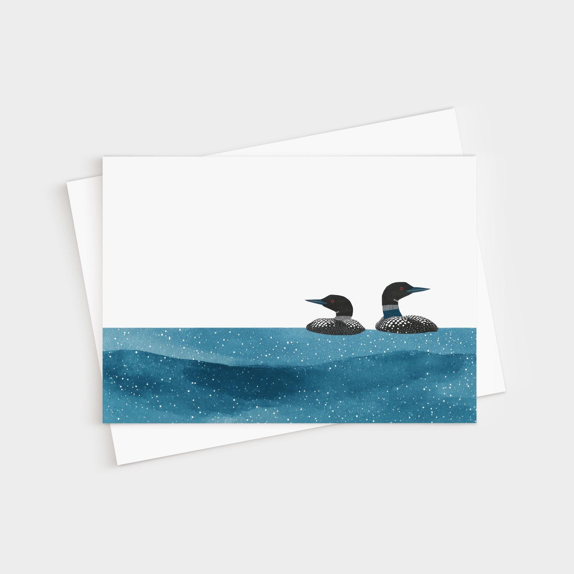 Minimalist greeting card with two loons illustrated in black and white, floating on a deep blue, star-speckled water pattern against a clean white background, paired with a white envelope.