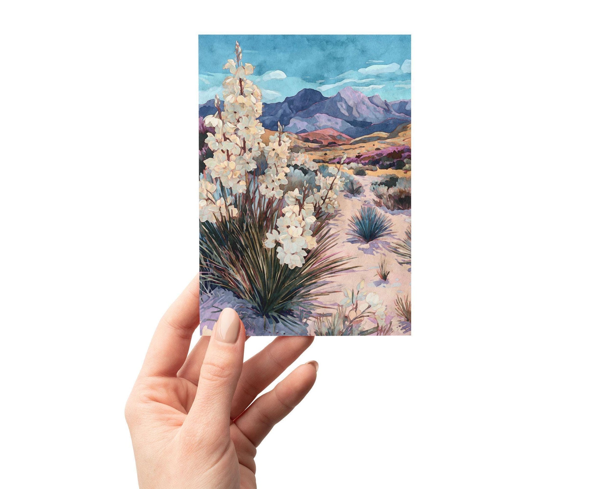 Hand holding a greeting card featuring a watercolor-style desert landscape with blooming yucca plants in the foreground, sandy terrain with scattered shrubs, and purple mountains under a vivid blue sky.