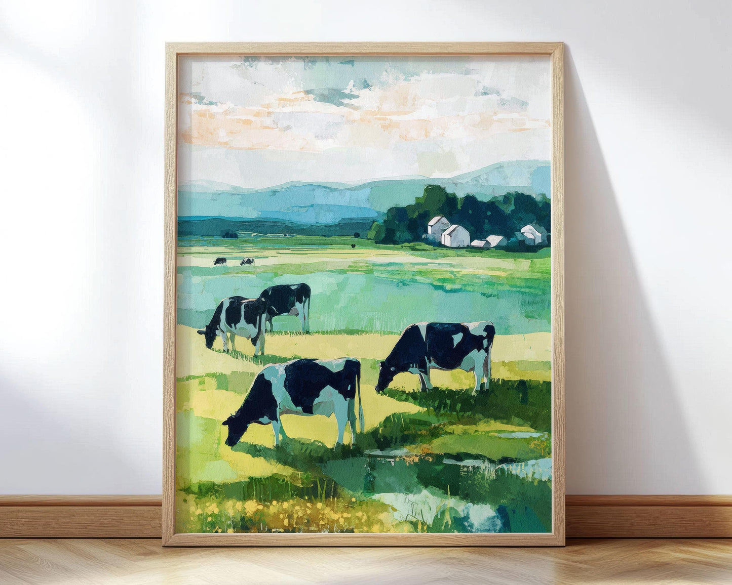 Framed painting of a pastoral countryside scene with black and white cows grazing in a green meadow. In the distance, a cluster of white farm buildings sits near a line of trees, with rolling blue hills under a partly cloudy sky. The artwork uses bold, painterly brushstrokes in greens, blues, and warm neutrals for a serene, modern impressionist look.