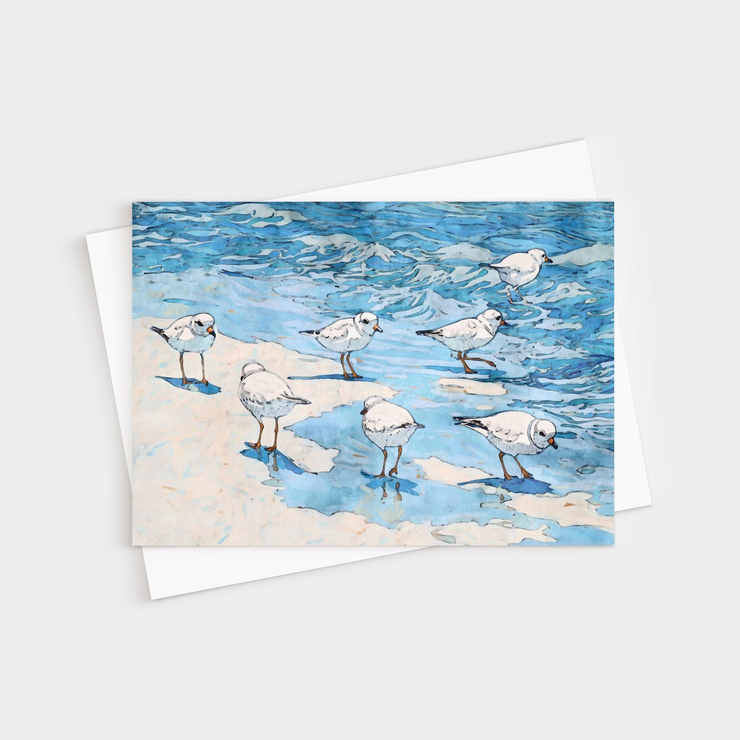 Watercolor-style illustration of small white shorebirds with orange legs standing and wading at the edge of blue ocean waves on a sandy beach, printed on a folded greeting card with a white envelope behind it.