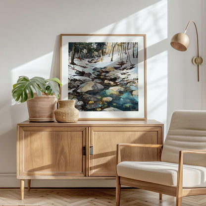 Framed watercolor-style painting of a snowy forest stream with rocks and reflections displayed on a wooden sideboard. The scene is styled with a potted monstera plant, a woven vase, and a modern beige armchair nearby. Sunlight streams through the window, casting soft shadows, creating a warm, minimalist, Scandinavian-inspired interior.