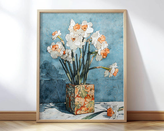 Framed art print of white daffodils with orange centers in a patterned floral vase against a textured blue background, displayed leaning against a wall on a wooden floor.