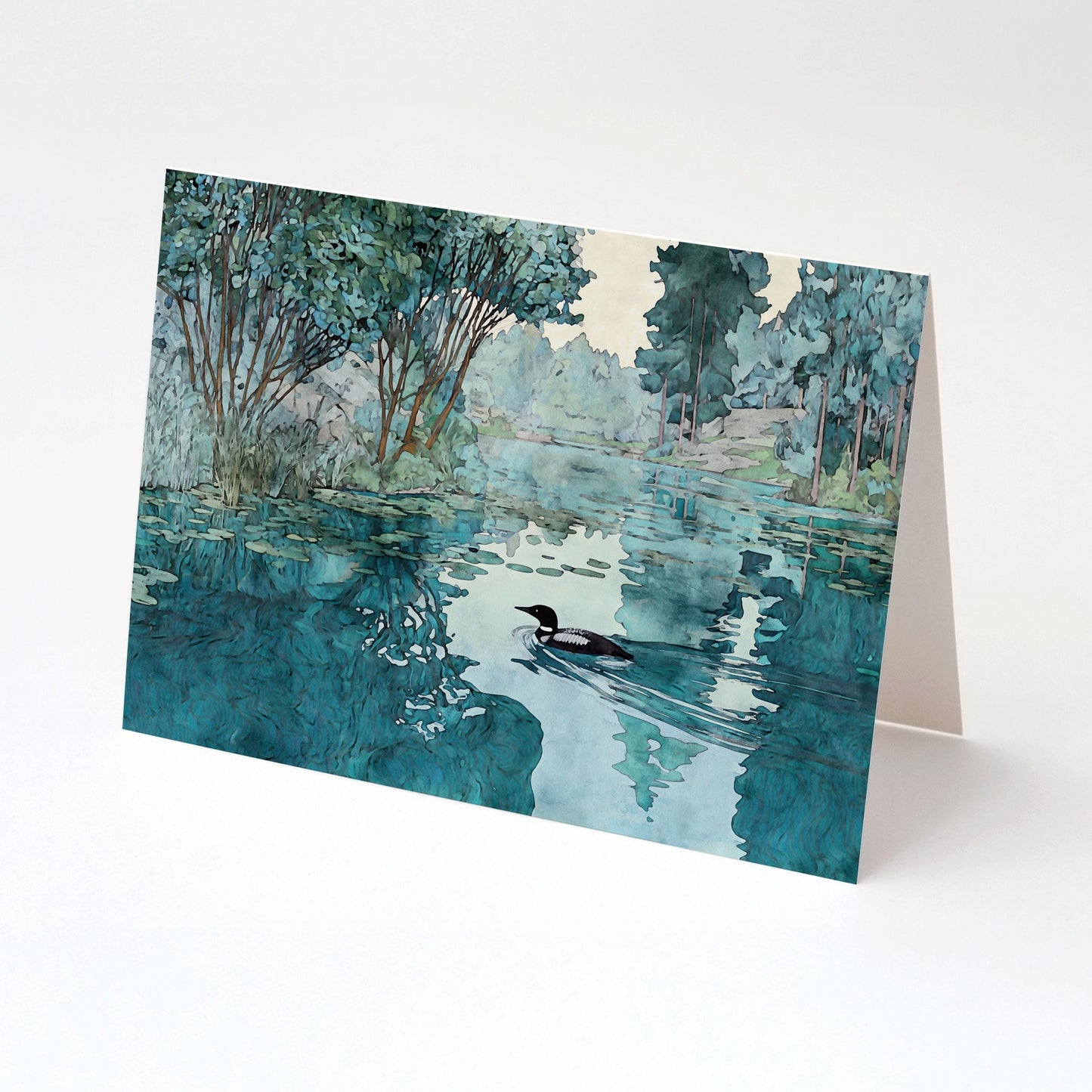 Folded greeting card featuring a watercolor-style illustration of a loon gliding across a reflective lake with trees and greenery along the shore. The card is displayed standing against a clean white background.