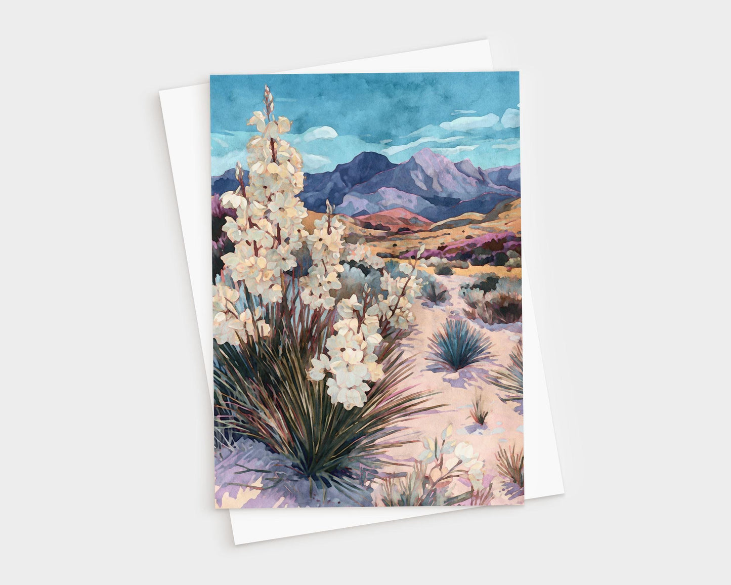 Greeting card featuring a watercolor-style desert landscape with blooming yucca plants in the foreground, sandy terrain with scattered shrubs, and purple mountains under a vivid blue sky, paired with a white envelope.