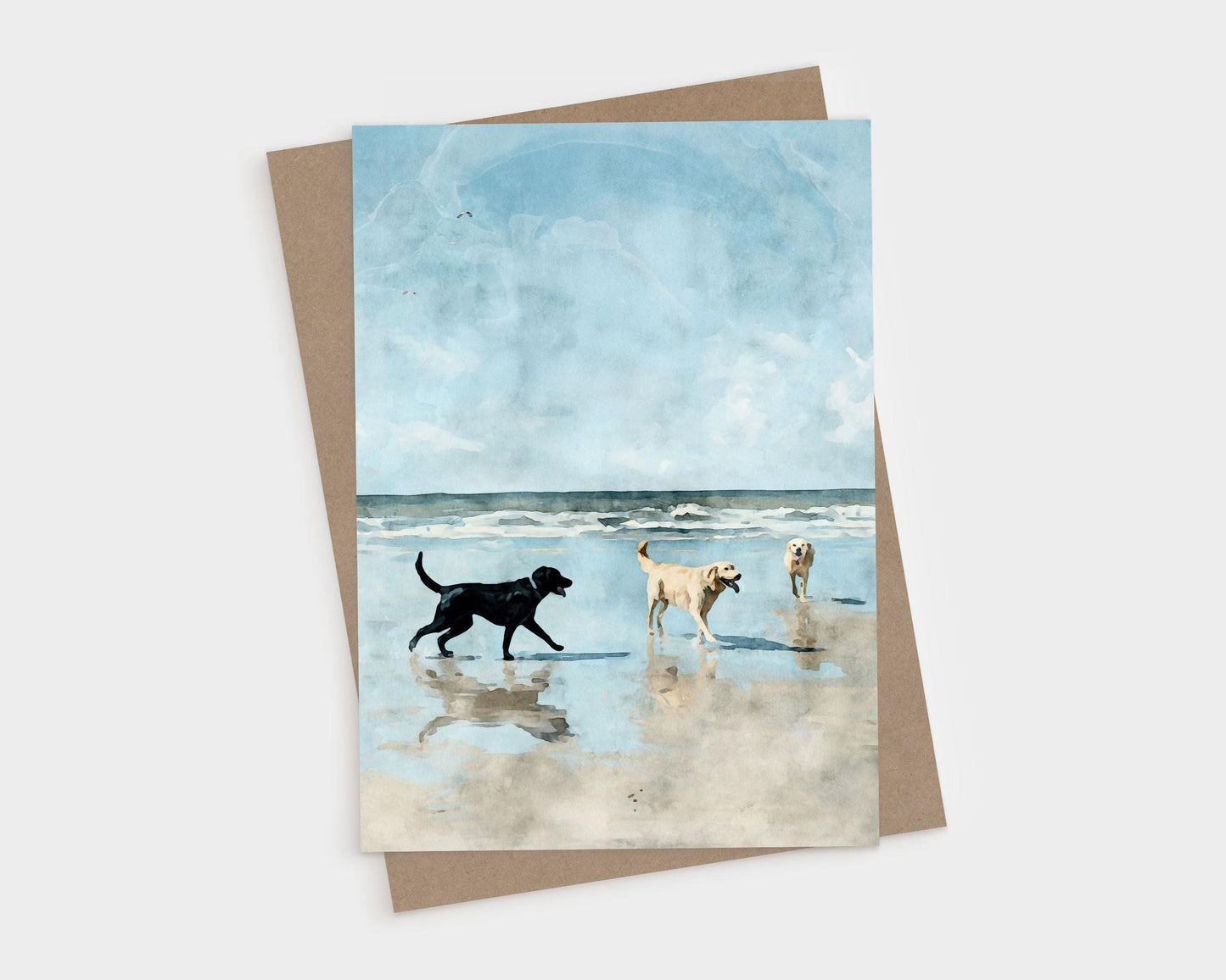 Watercolor-style illustration of three dogs—one black and two golden—playing on a sandy beach with gentle waves and a bright blue sky in the background, printed on a greeting card with a kraft paper envelope behind it.