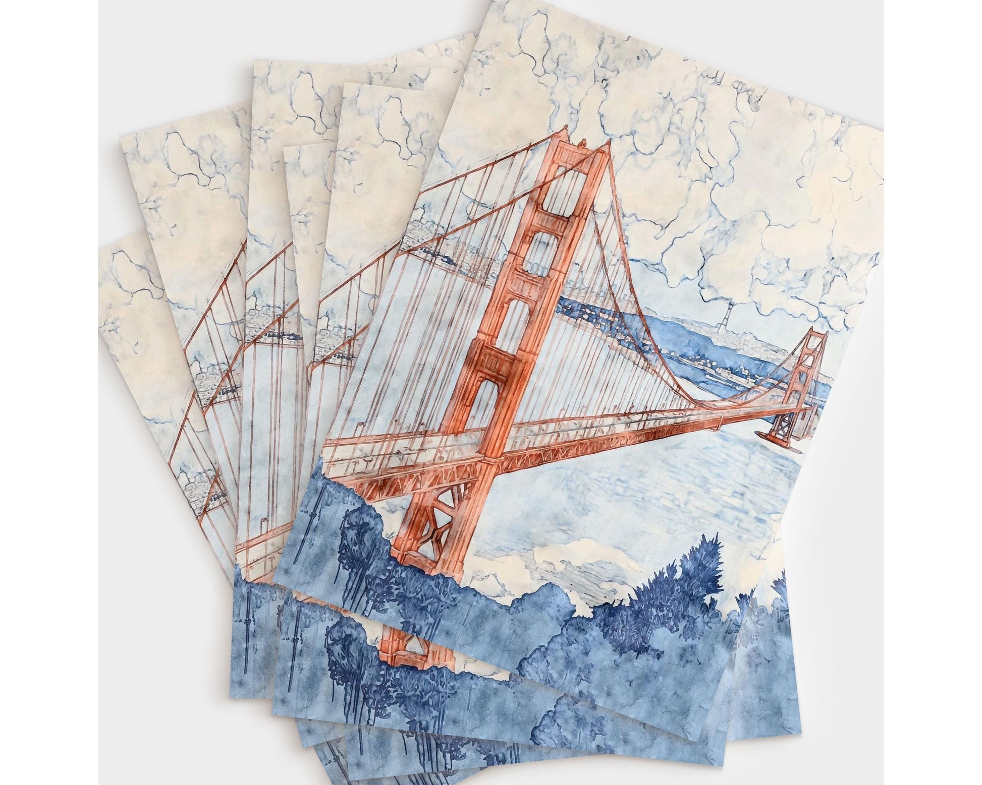 Flat lay of six greeting cards featuring a watercolor painting of the Golden Gate Bridge in warm red-orange tones, set against a cloudy sky and soft blue bay. The foreground includes bold navy foliage, creating contrast with the pastel sky and water.