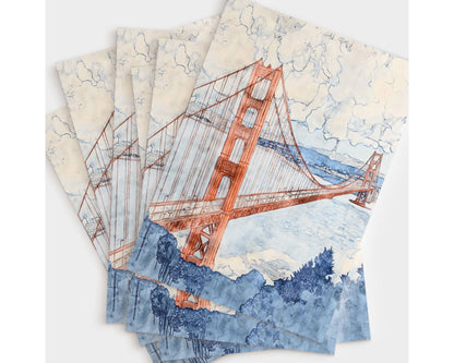 Flat lay of six greeting cards featuring a watercolor painting of the Golden Gate Bridge in warm red-orange tones, set against a cloudy sky and soft blue bay. The foreground includes bold navy foliage, creating contrast with the pastel sky and water.