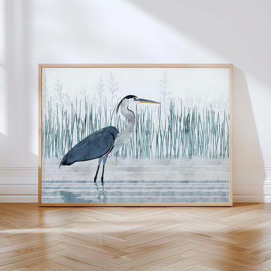 Framed art print of a stylized great blue heron standing in shallow water, with tall marsh grasses in muted green and gray tones behind it. The heron has a sleek, minimalist form with navy and gray plumage, and the scene is rendered in a soft, watercolor-inspired style. The artwork is propped against a white wall on a light wood herringbone floor, with gentle natural light casting soft shadows.