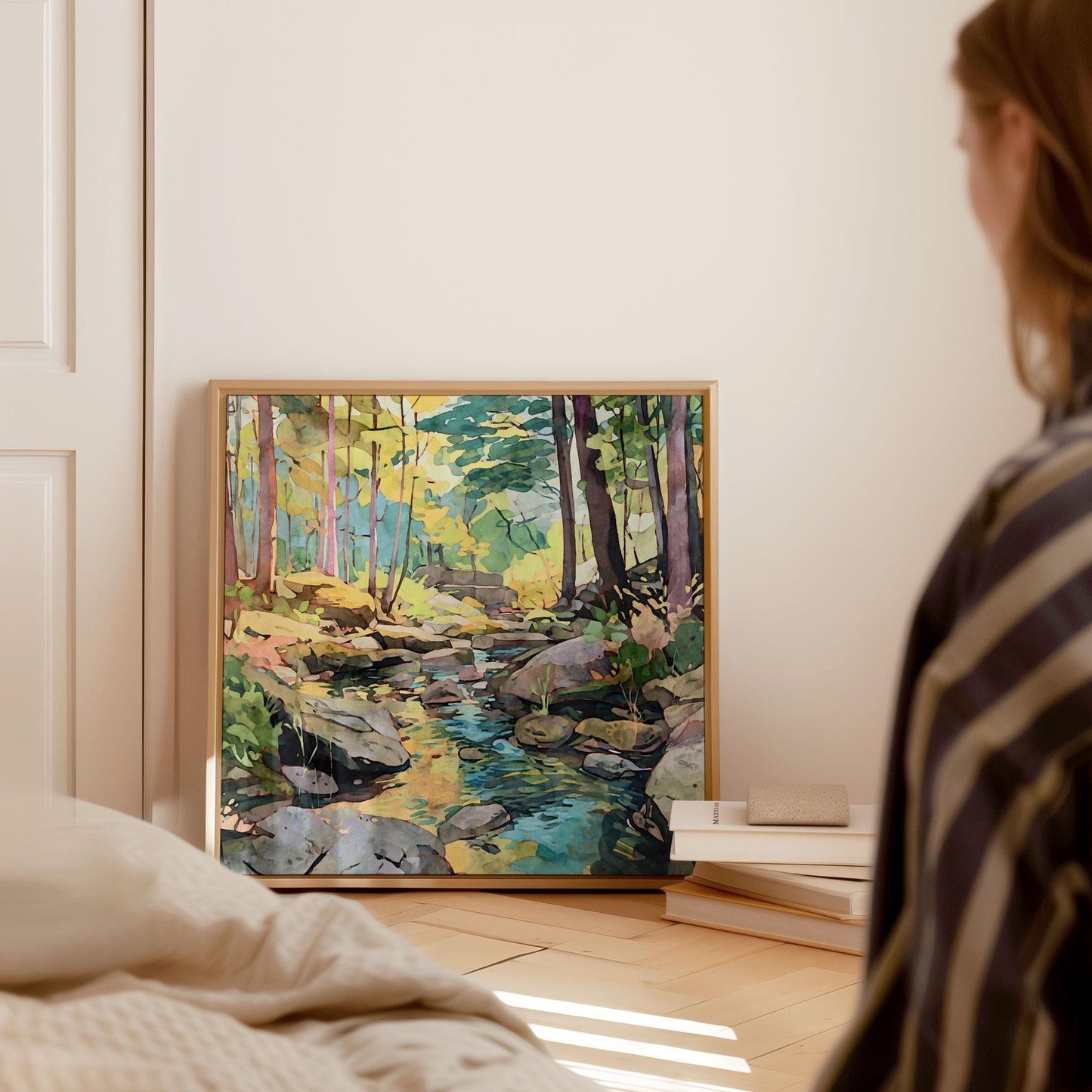 Framed watercolor-style forest stream painting rests on the floor against a wall, beside a stack of books, with a person in a striped shirt seated nearby in a softly lit, cozy bedroom setting.