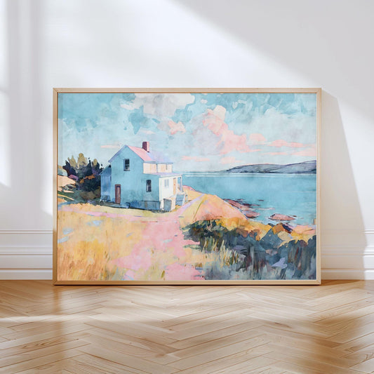 Framed watercolor-style painting of a coastal scene showing a light blue house on a grassy bluff overlooking the ocean. The sky is filled with soft pastel clouds in pinks and blues, and the path leading to the house is painted in warm peach and yellow tones. The artwork rests against a white wall on a herringbone wood floor, bathed in natural light.
