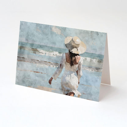 Greeting card displayed upright on a clean white background, featuring a watercolor-style image of a woman in a white dress and wide-brimmed hat walking toward the ocean waves under a soft blue sky.
