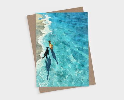 Watercolor-style greeting card featuring two dogs walking along the shoreline, their shadows stretching across the shallow turquoise waves. The sandy beach contrasts with the vivid blue water, creating a peaceful, sunlit scene. A kraft envelope is partially visible behind the card.