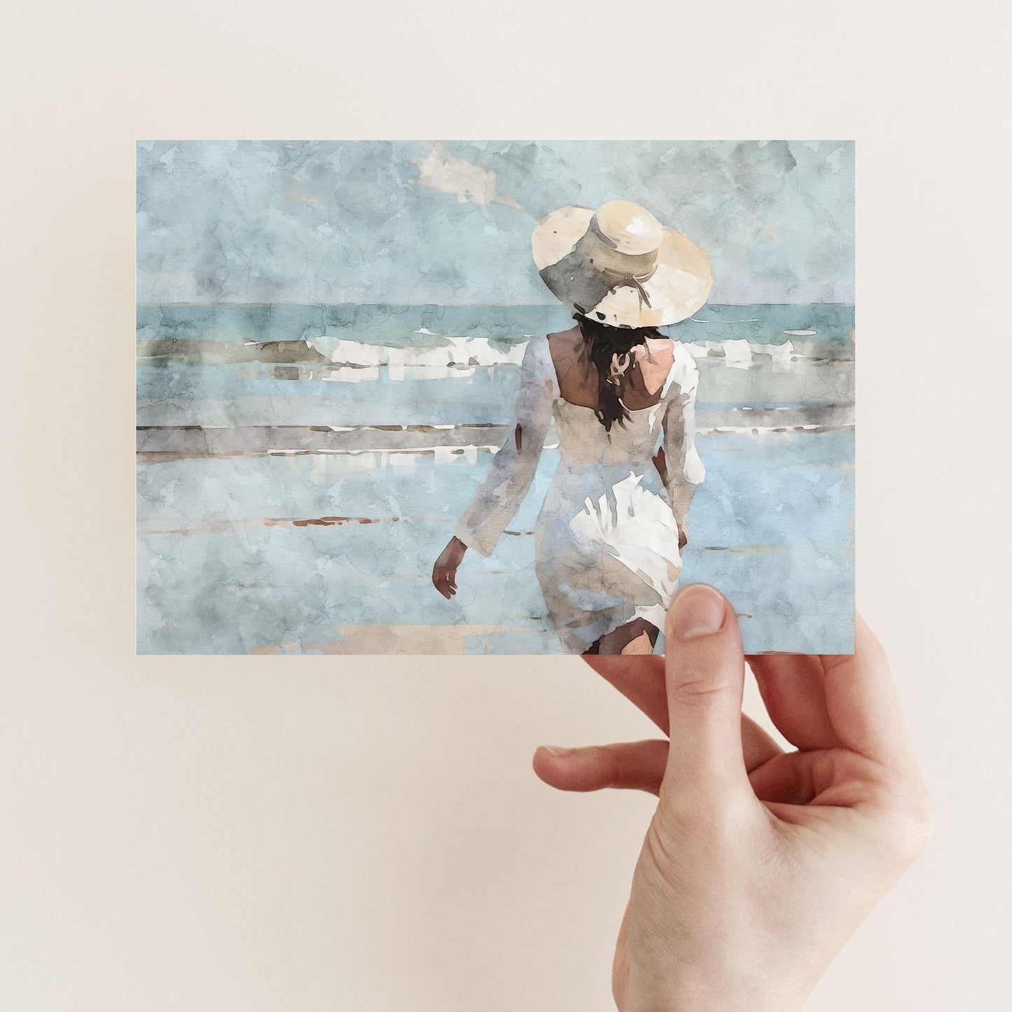 A hand holds a watercolor-style greeting card depicting a woman in a white dress and sunhat walking toward gentle ocean waves under a soft blue sky.