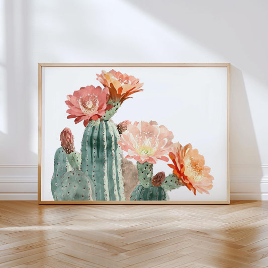 Watercolor art print of a blooming cactus with large coral, peach, and orange flowers, set in a light wood frame and leaning against a white wall on a herringbone wood floor.