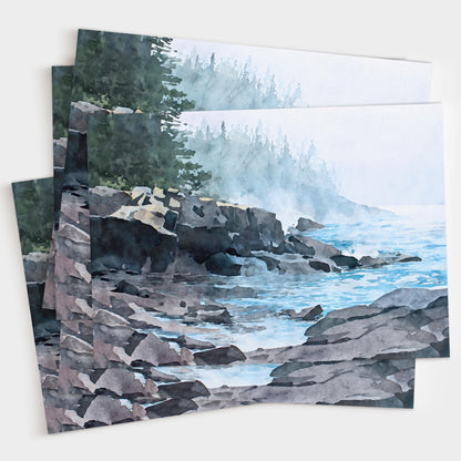 Set of horizontal greeting cards fanned out on a light background, each featuring the same watercolor-style illustration of a misty Lake Superior shoreline. Dark rocky cliffs and layered stones meet calm blue water with gentle ripples. Pale fog softens the horizon and partially obscures a distant evergreen forest, creating a quiet, atmospheric lakeside scene in muted blues, grays, and greens.