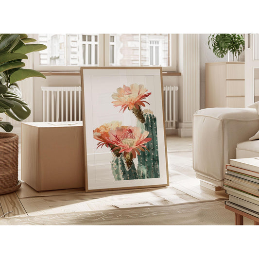 Framed watercolor cactus print with peach and coral flowers leaning against a box in a bright, modern living room with neutral furniture, wood floors, and potted plants.