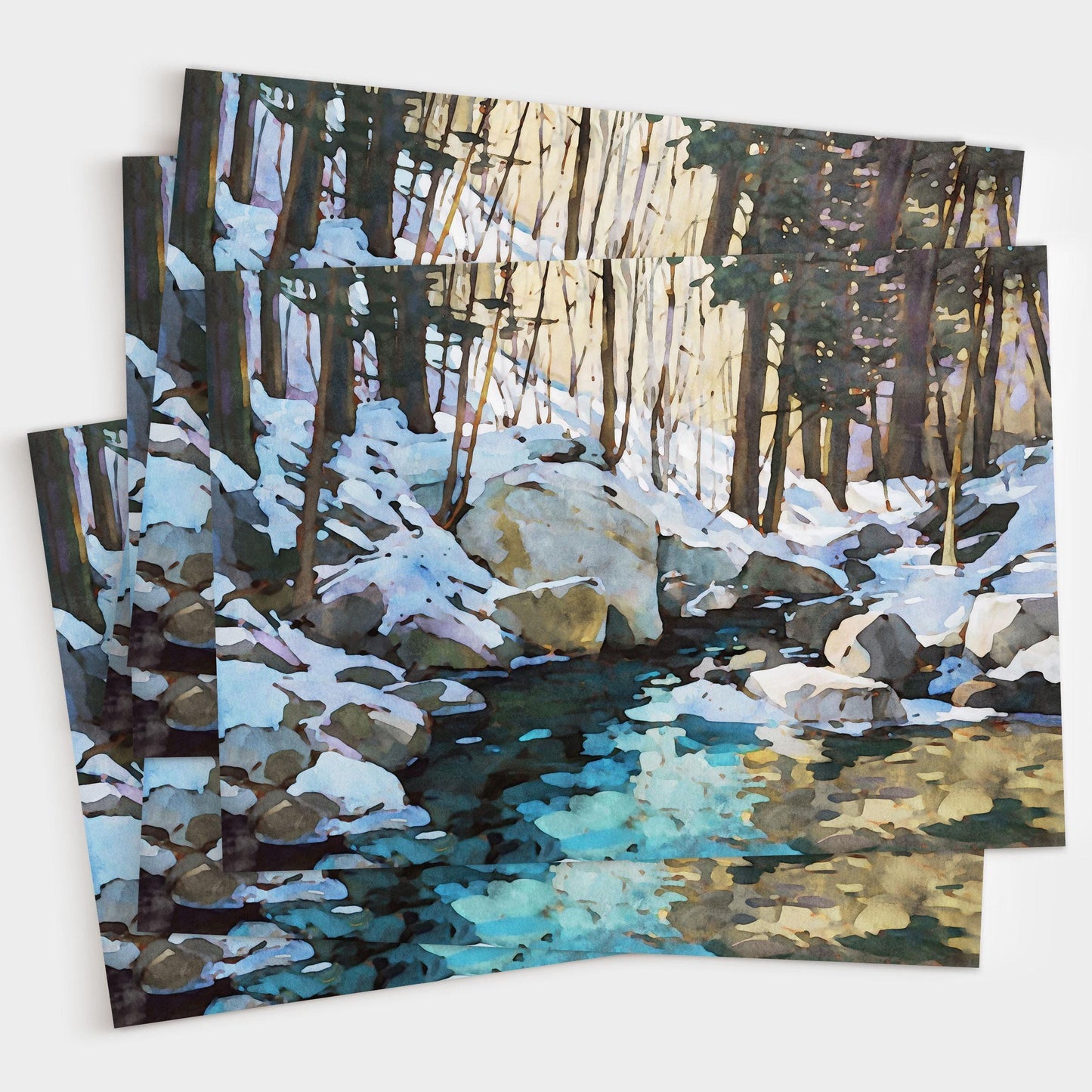 Set of greeting cards featuring a watercolor-style snowy forest scene with large rocks and a creek reflecting icy blue and golden tones, shown stacked and slightly fanned out to display multiple copies.