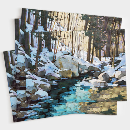 Set of greeting cards featuring a watercolor-style snowy forest scene with large rocks and a creek reflecting icy blue and golden tones, shown stacked and slightly fanned out to display multiple copies.