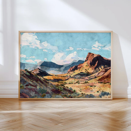 Framed painting of a sunlit desert canyon scene with rugged red rock cliffs, rolling hills, and distant blue mountains under a bright sky filled with fluffy white clouds, leaning against a white wall on a wooden herringbone floor.