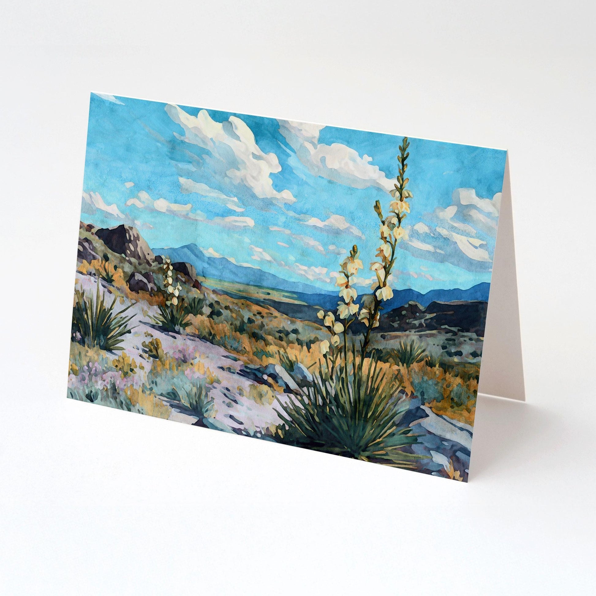 Greeting card standing open, featuring a watercolor-style desert landscape with blooming yucca plants, rocky hills, and distant blue mountains under a vivid sky filled with white clouds.