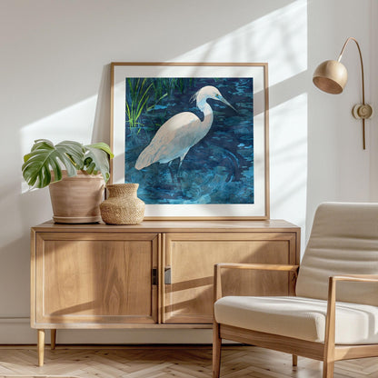 Framed watercolor art print of a white egret standing in rippling blue water with green reeds, displayed on a wooden sideboard beside a potted plant and woven vase in a sunlit modern living room with a neutral armchair and wall-mounted lamp.