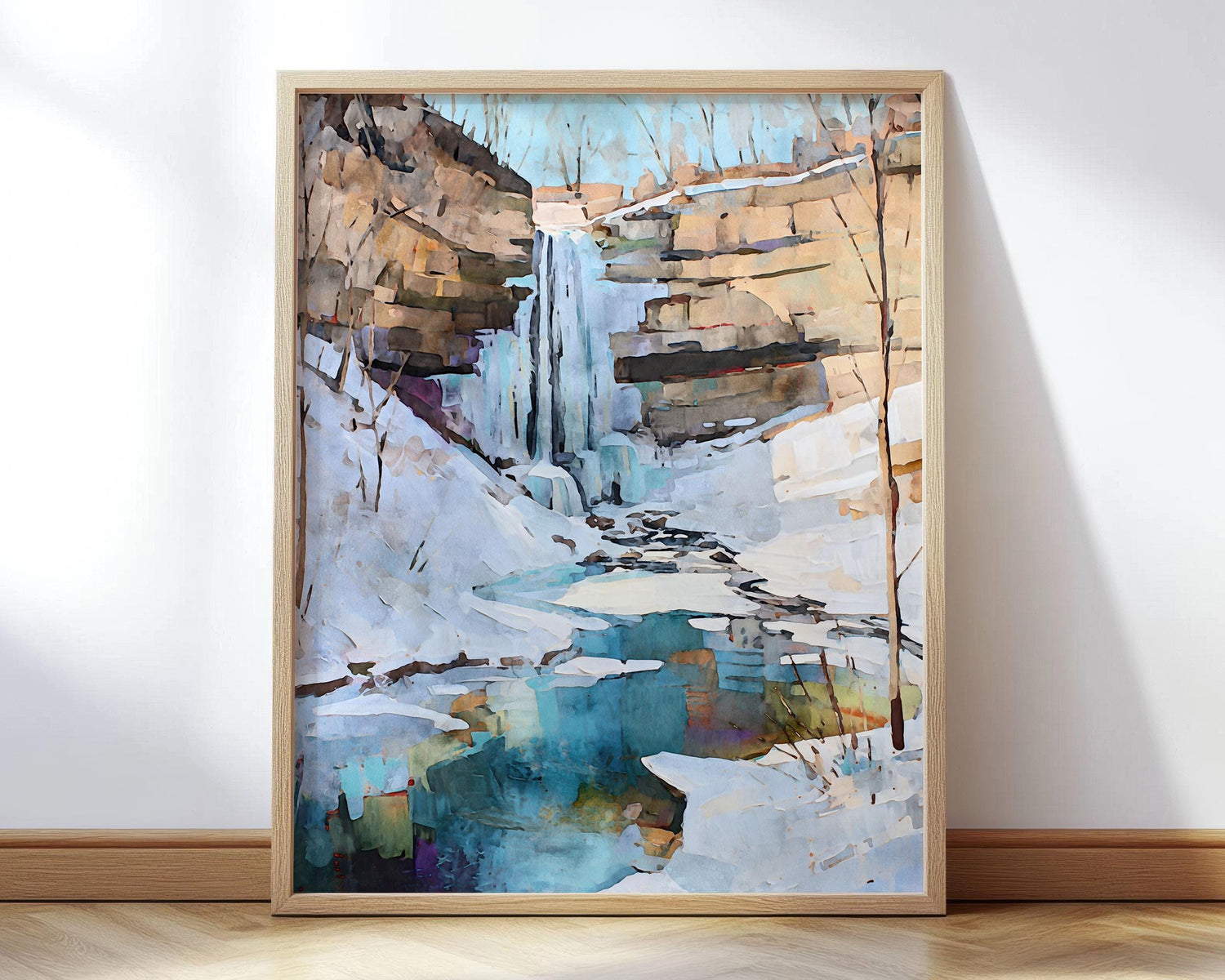Framed watercolor-style painting of a winter waterfall scene, showing icy cliffs, snow-covered ground, and a turquoise pool below, leaning against a white wall on a wooden floor.
