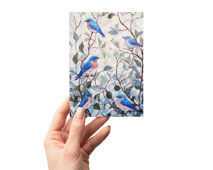 Hand holding a greeting card featuring a watercolor design of five bluebirds with peach-colored chests perched among soft green and brown branches against a pale, dappled background.