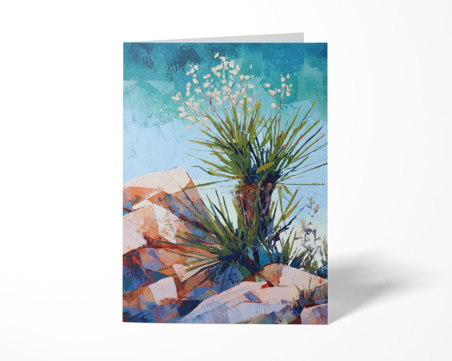 Standing greeting card with a colorful desert landscape painting of a yucca plant, its spiky green leaves and white blossoms set against a bright blue sky and warm rocky terrain in shades of red, orange, and purple.