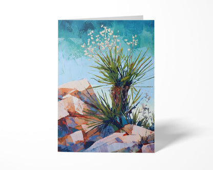 Standing greeting card with a colorful desert landscape painting of a yucca plant, its spiky green leaves and white blossoms set against a bright blue sky and warm rocky terrain in shades of red, orange, and purple.