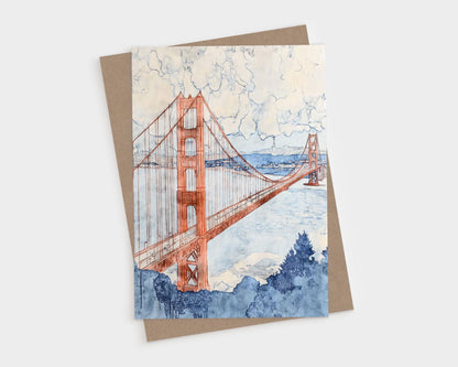 Greeting card featuring a watercolor illustration of the Golden Gate Bridge in warm terra-cotta tones, stretching across a pale blue bay. Billowing clouds fill the sky above, with soft washes of navy and indigo detailing the foliage in the foreground and the San Francisco skyline in the distance.
