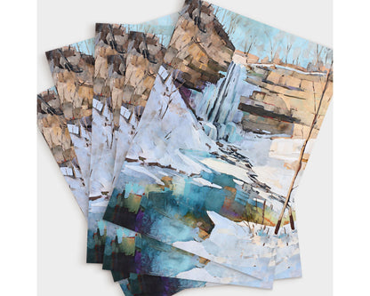 Set of greeting cards displayed in a fanned arrangement, each featuring a watercolor-style painting of a snowy winter waterfall scene. The artwork shows a cascade flowing over rocky cliffs into a turquoise pool, surrounded by snow and bare trees under a pale sky.