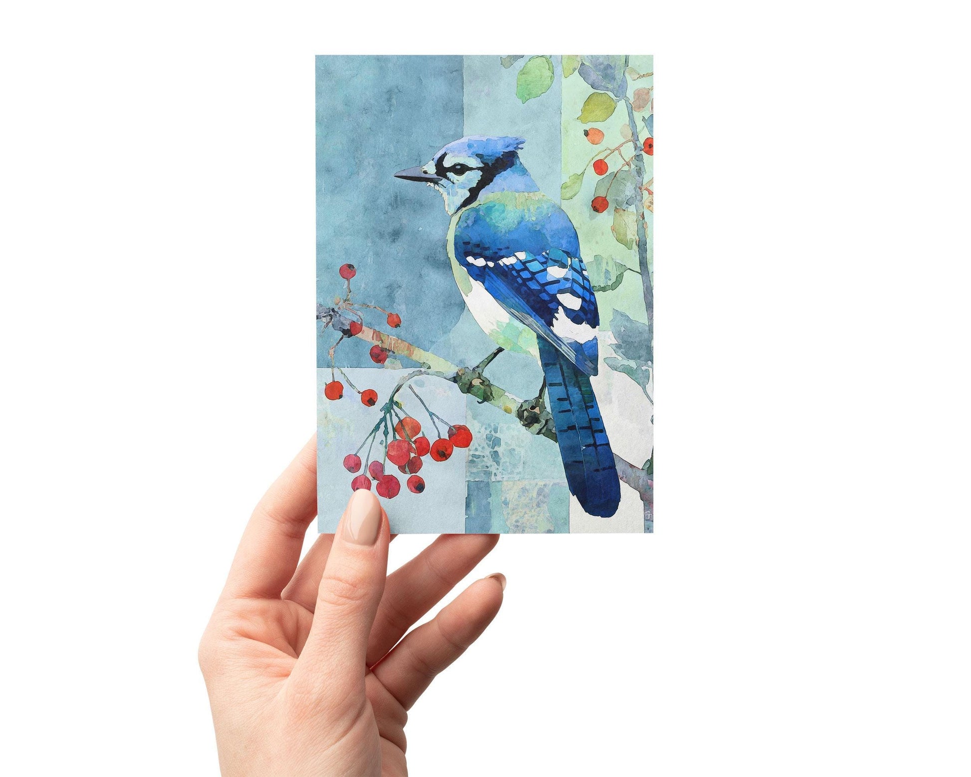 Hand holding a greeting card featuring a watercolor painting of a blue jay perched on a branch with red berries, set against a softly textured blue and green patchwork background. The artwork combines delicate natural detail with an abstract, modern feel.