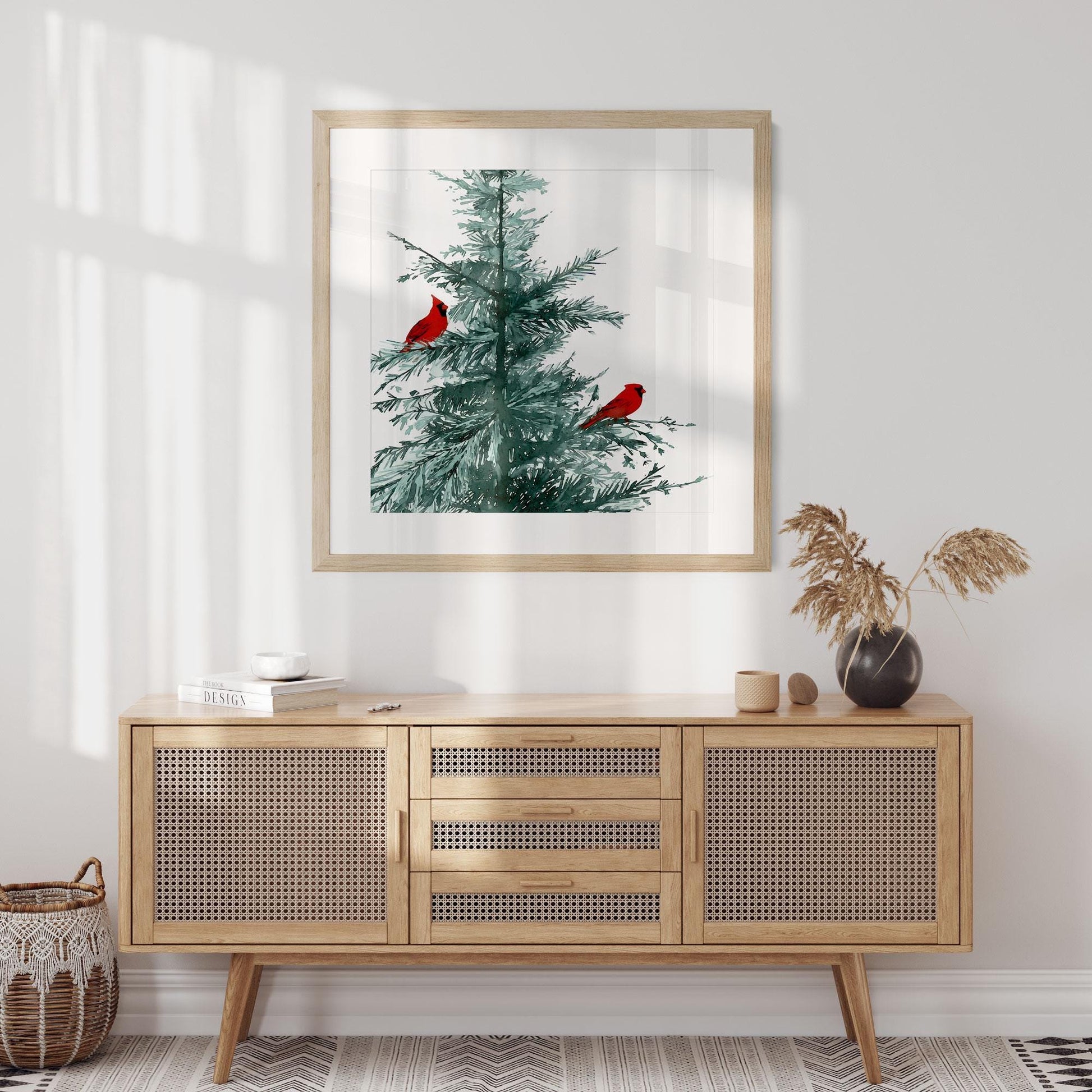 Framed watercolor art print of a pine tree with two bright red cardinals, hanging above a mid-century rattan sideboard in a light-filled modern room with neutral tones and minimalist decor.